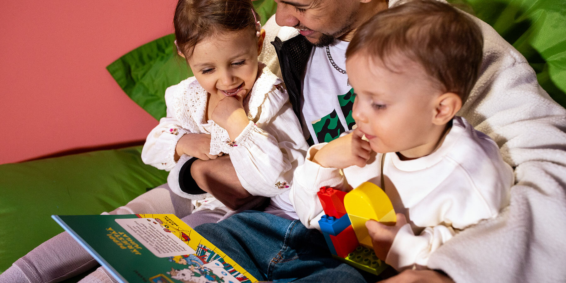 Kaksi pientä lasta lukee kuvakirjaa yhdessä aikuisen kanssa. Lapset istuvat aikuisen sylissä.