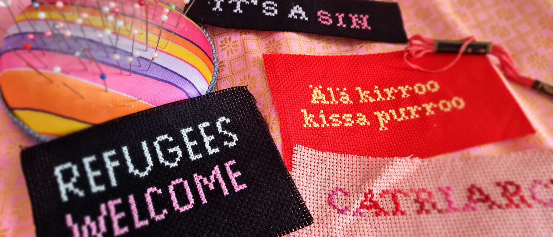 Erilaisia ristipistotöitä vierekkäin. Niissä on tekstejä: refugees welcome, it's a sin, älä kirroo - kissa purroo. Töiden vieressä on värikäs neulatyyny.