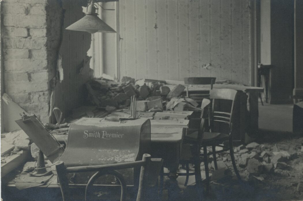 Taisteluissa vahingoittunut toimistohuone, tiilimurskaa lattialla, pöydällä Smith-kirjoituskone.