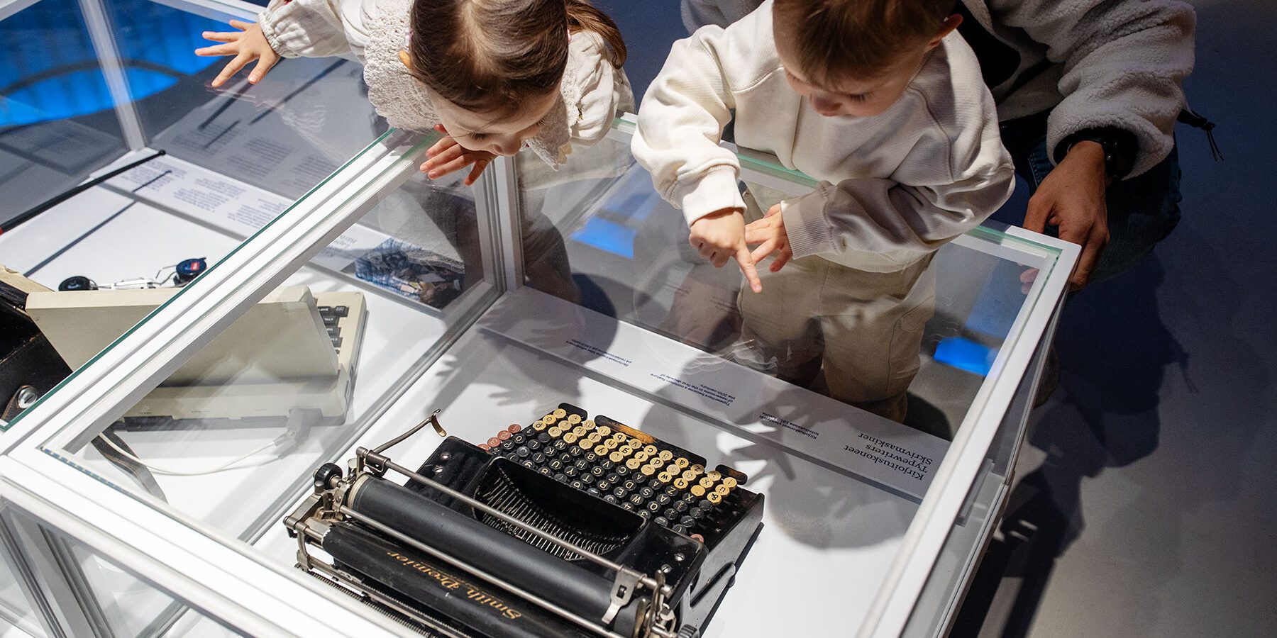 Kaksi lasta tutkii museon esinevitriinissä olevaa kirjoituskonetta aikuisen kanssa.